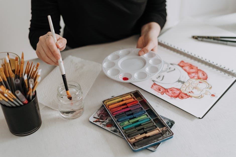 Unrecognizable painter with palette and paintbrush sitting at table with sketchbook and art supplies while painting in modern light studio